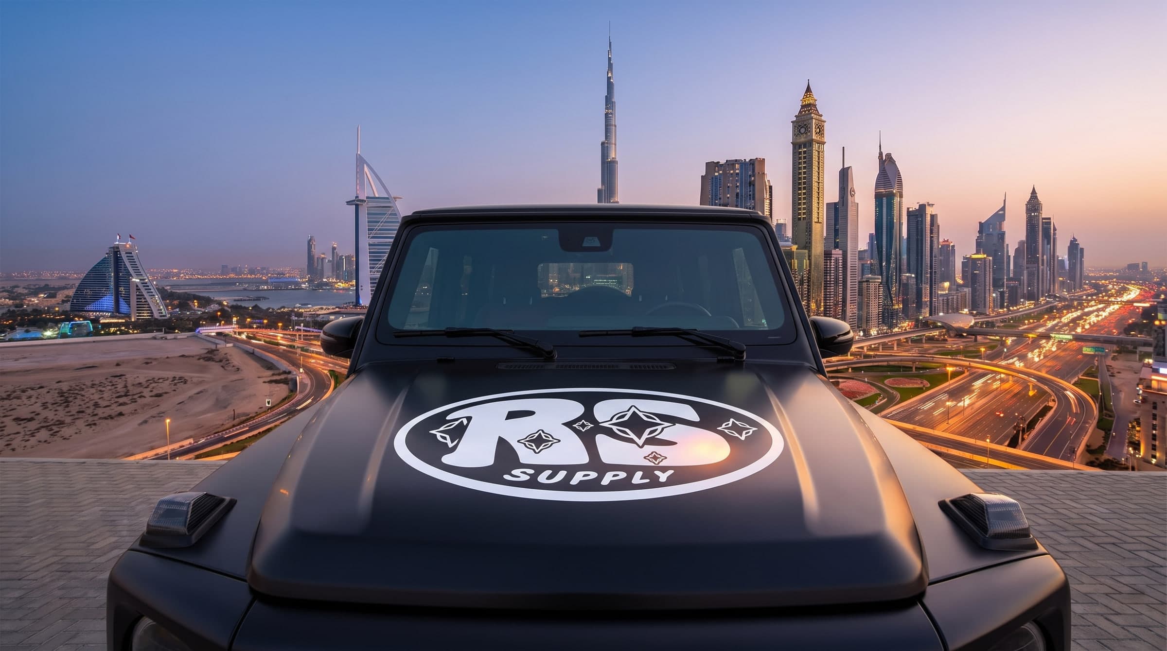 The RS Supply oval on a matte black hood, Dubai skyline behind it at dusk